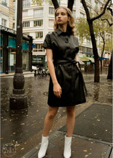 Woman in a black leather dress standing on a city street with buildings and trees in the background.